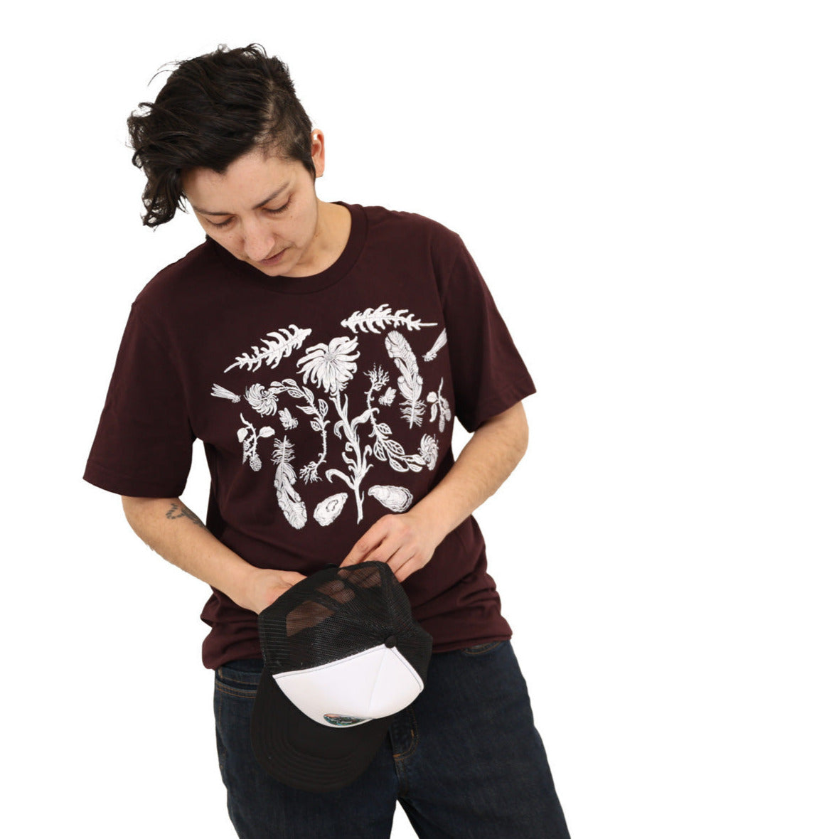 Woman wearing a oxblood colored t-shirt with white ink of flowers, ferns, feathers, shells, etc