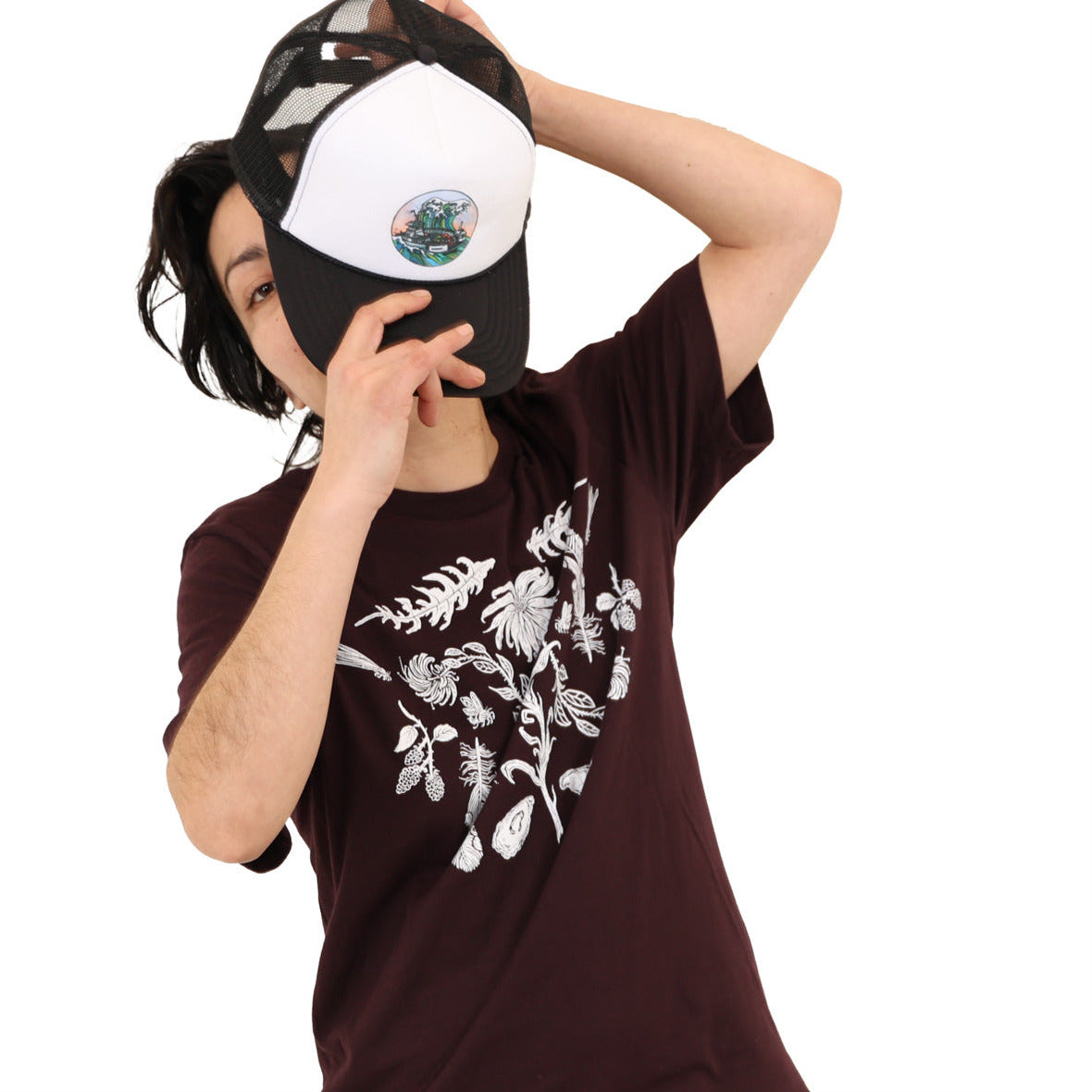 Woman wearing a oxblood colored t-shirt with white ink of flowers, ferns, feathers, shells, etc. Woman has hat half way over her face