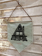 Green canvas pennant printed with black A-Frame house design, hanging on wall 3/4 view.