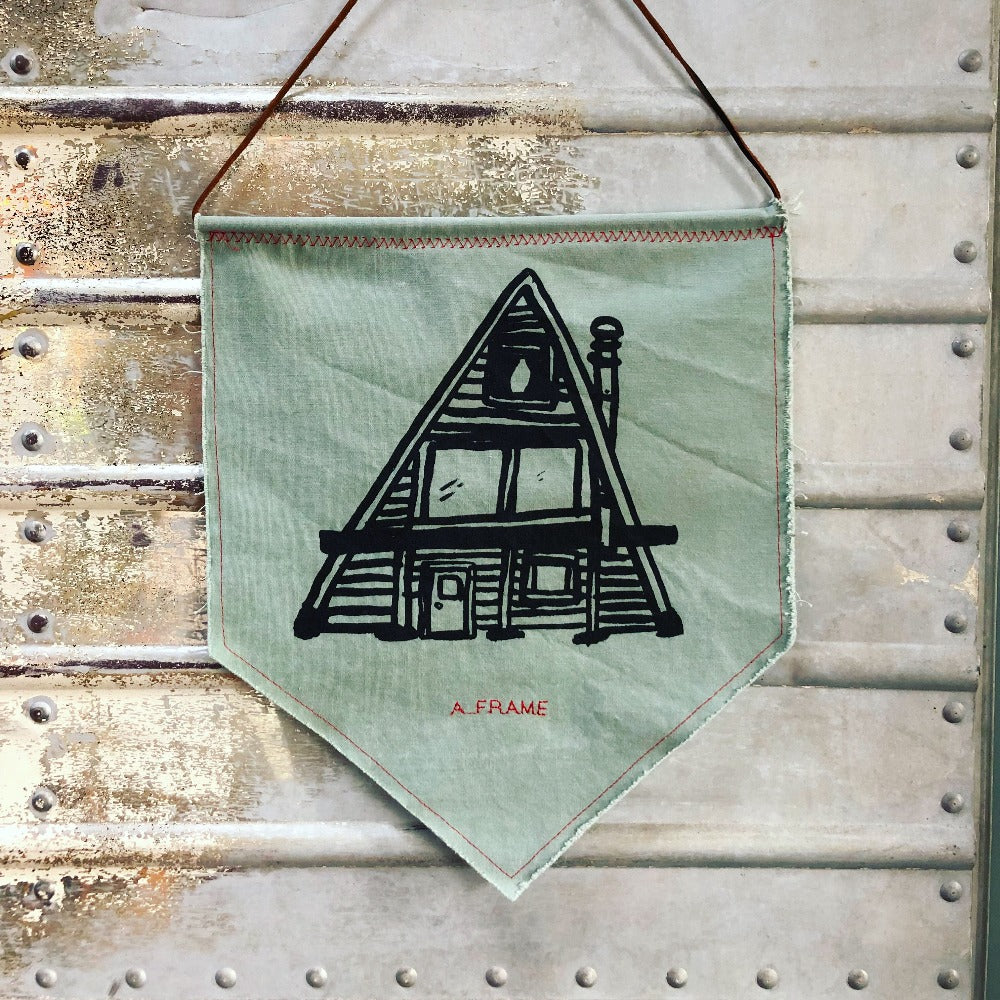Green canvas pennant printed with black A-Frame house design, hanging on wall.
