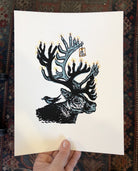 Art print in hand - Painting of caribou with candles for antlers and small tag reading, "be the light". Magpie perched on caribou's back.