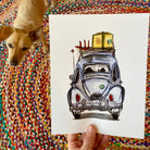 The Dog Is My Copilot art print, a colorful painting of a VW bug from the rear, with silhouettes of a long-haired person in the driver's seat and a dog in the passenger seat. The print is in the artist's hand, in front of a braided rug and the artist's cutie-pie dog, Cricket.