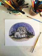 Colorful art print of a geodesic dome house in the desert, under a purple night sky. Watercolor and ink. Laid on the artist's desk next to brushes and pens.
