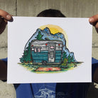 Colorful print - a painting of a vintage Bell camper trailer in a mountainside camp. Print is being held up by the corners, for scale.