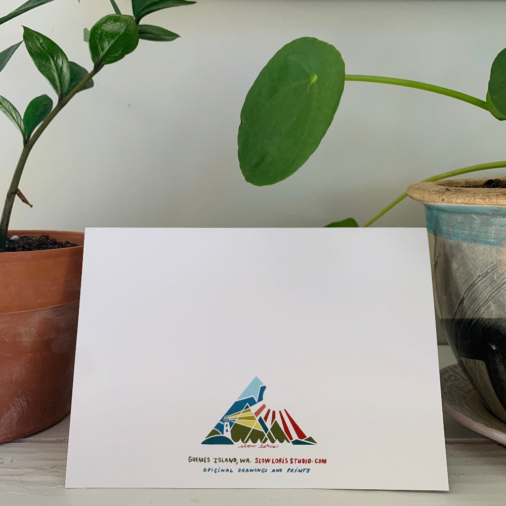 The back of a folded greeting card sitting on a desk with potted plants. The back is printed with a logo and information for Slow Loris , the card's brand.