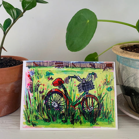 A greeting card sitting on a table with potted plants. The card features a colorful painting of a bicycle in a field full of flowers.