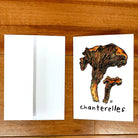 A greeting card printed with two colorful chanterelles, lying on a table next to a white envelope.