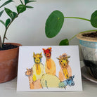 A greeting card, printed with a colorful tissue and ink painting of golden alpacas. The card sits on a table between two plants.