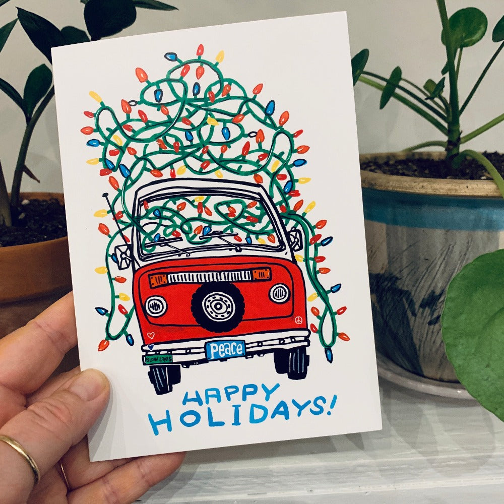 A hand holding a greeting card printed with a colorful painting of a van bedecked in holiday lights, with a license plate reading "peace" and the words Happy Holidays in hand lettering below.
