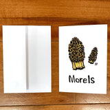 A greeting card printed with a painting of morel mushrooms and the word "morels" in hand-lettering below. The card lies flat on a table next to an envelope.
