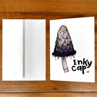 A greeting card printed with a painting of an inky cap mushroom and the words Inky Cap in hand-lettering. The card is lying flat on a table next to an envelope.