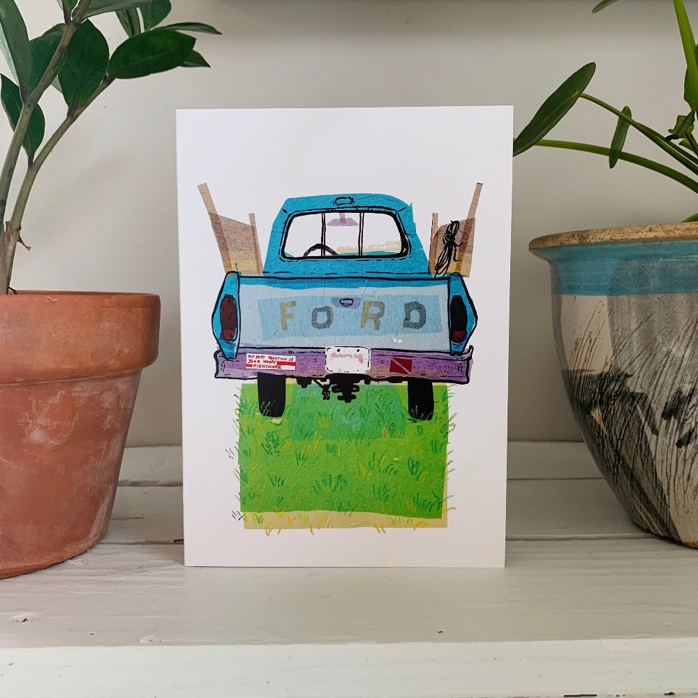 A greeting card printed with a colorful ink and tissue painting of an old blue Ford truck on a patch of grass. The card is on a table between two houseplants.