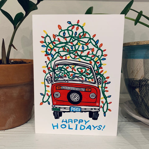 A greeting card printed with a colorful painting of a van bedecked in holiday lights, with a license plate reading "peace" and the words Happy Holidays in hand lettering below.