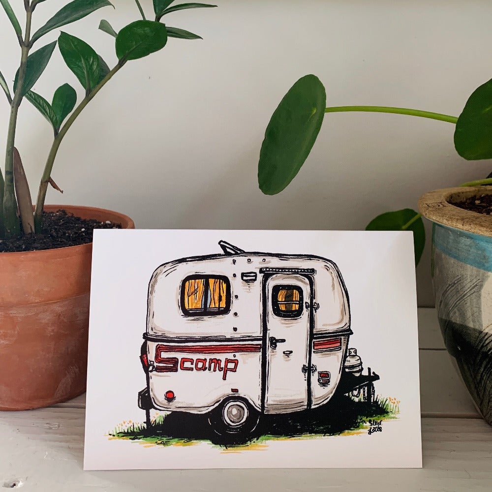 A greeting card printed with a colorful painting of a vintage Scamp trailer camper. The card sits on a table between two plants.