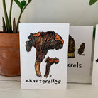 A greeting card printed with a colorful painting of a Chanterelle mushroom on the front, with the word Chanterelle in hand-lettering below. In the background are a greeting card with Morel mushrooms and one with an Inky Cap mushroom done in the same style.
