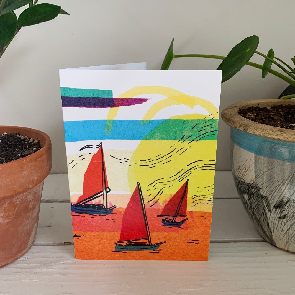 Greeting card printed with a colorful ink and tissue painting of three boats setting sail at sunset. The card is on a table between two plants.