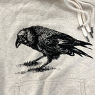 Close up detail of an oatmeal-colored hoodie, screen printed with a black crow.