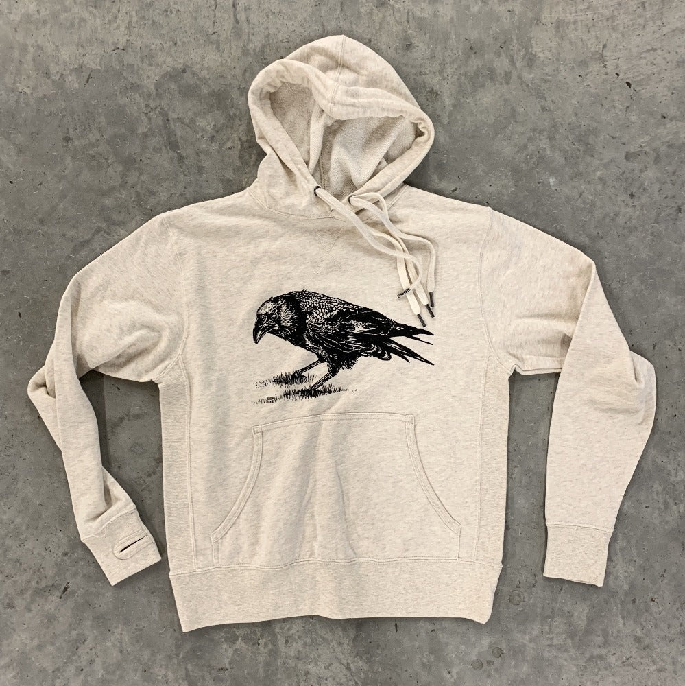 An oatmeal-colored hoodie, screen printed with a crow in black ink and laid flat against a gray background.