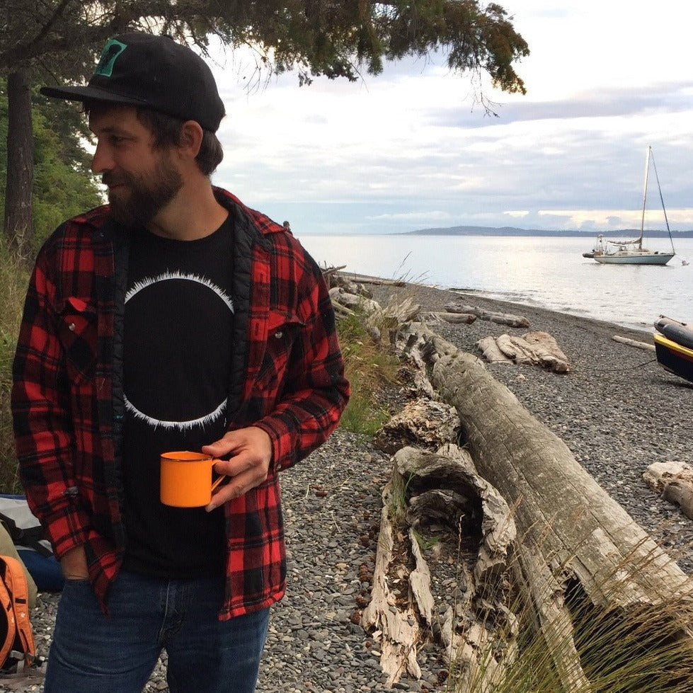 man on beach with sailboat in the background. Man is holding an orange cup talking with someone out of frame. Man is wearing a ball cap and a red flanel sweater that is open in the front showing the black t-shirt with white eclipse print on it. 