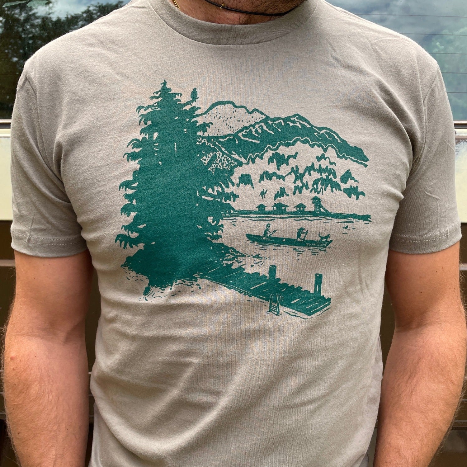 Man wearing a warm grey t-shirt with a green print of lakeside scene on it.