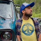 man leaning up against a Westfalia van while wearing a ball cap and t shirt with blue print on a yellow T. Print is of a campfire's smoke going up the center meeting with the tops of curved in trees creating a peace sign. Tent is by the fire and a bird is on a branch