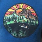 blue shirt with a multi-colored print of sunsetting behind the mountains, and a canoe lake camping scene