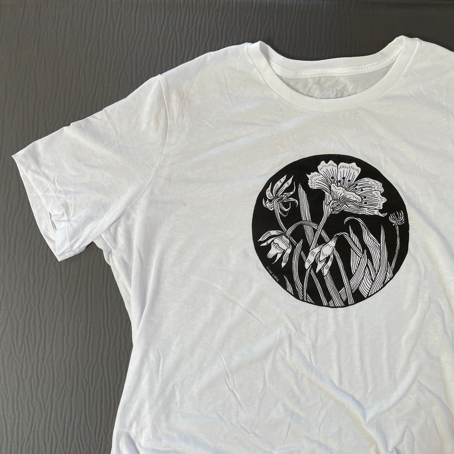 close up of white t shirt with black ink of an evening star flower and leaves framed in a circle. The background of the image is black and the flower line is white(the color of the shirt)