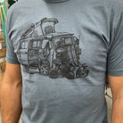 close up of man wearing grey blue shirt with a VW van open and top popped. Skateboards, bikes, and clothing strewn about. 
