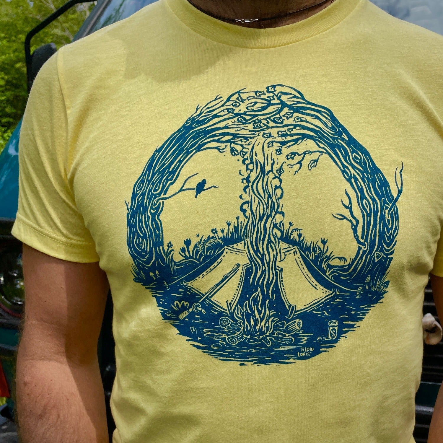 Close up of blue print on a yellow T. Print is of a campfire's smoke going up the center meeting with the tops of curved in trees creating a peace sign. Tent is by the fire and a bird is on a branch