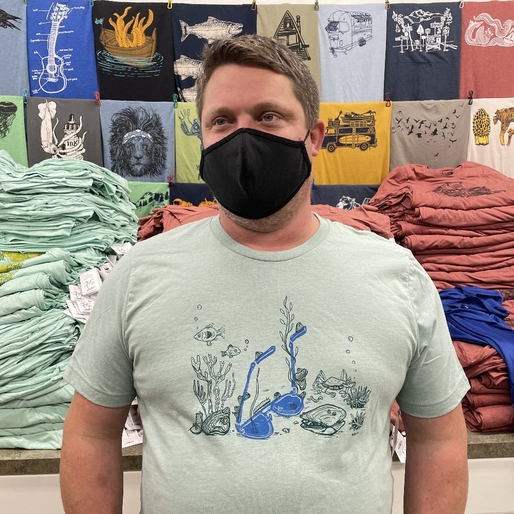 Man standing while wearing a mask on his face. Stacks of t-shirts behind. Man is wearing light soft blue t-shirt with print of sunglasses and sea creatures underwater