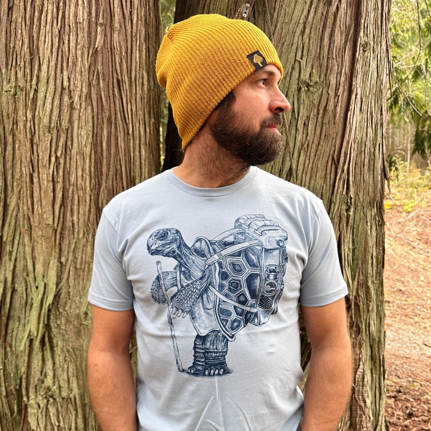 man standing in the forest wearing a mustard colored beanie and a blue t-shirt with a hiking tortoise in dark blue ink