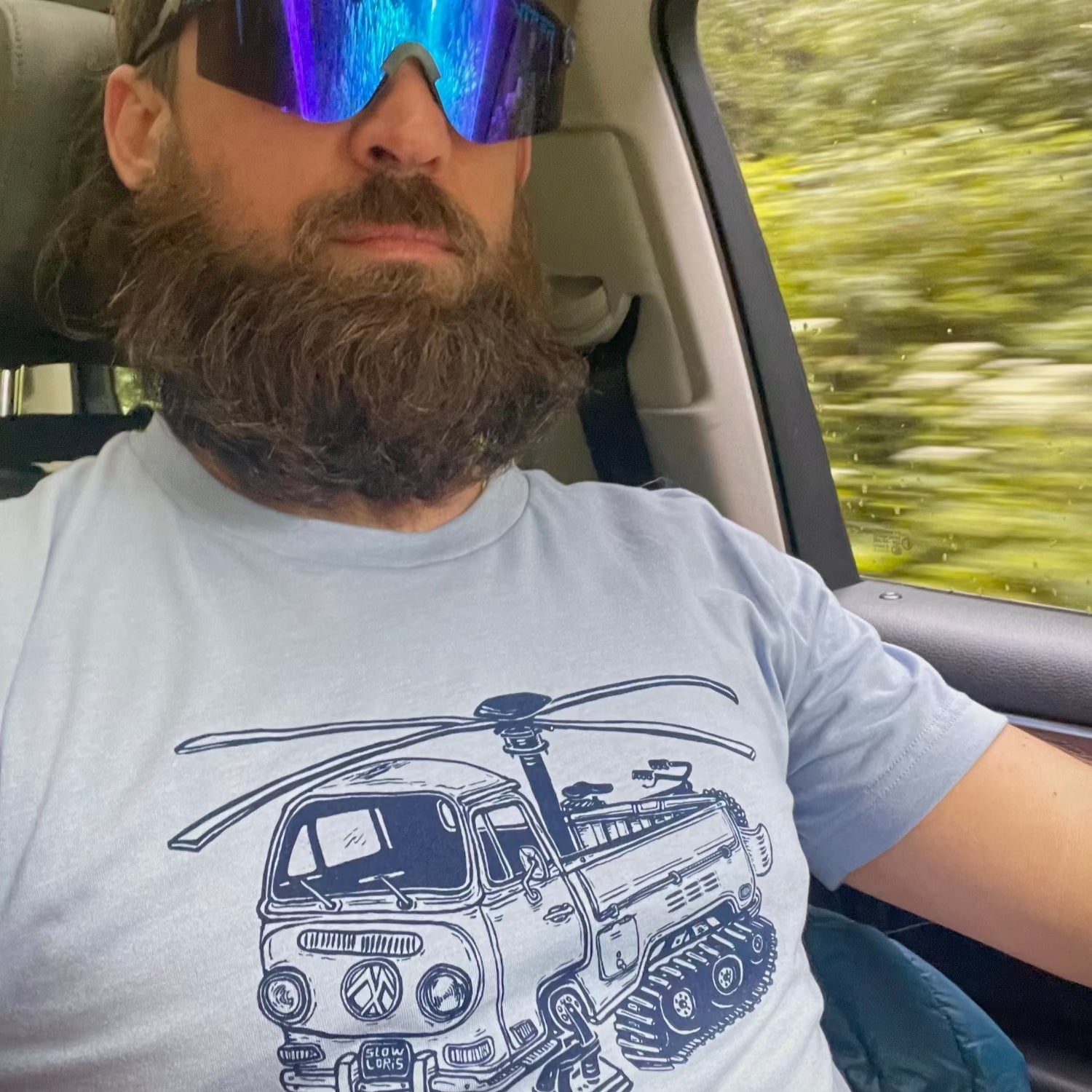 Man driving with a big beard and huge sunglasses while whering a shirt with the helicopter van with tracks and skis on it. 