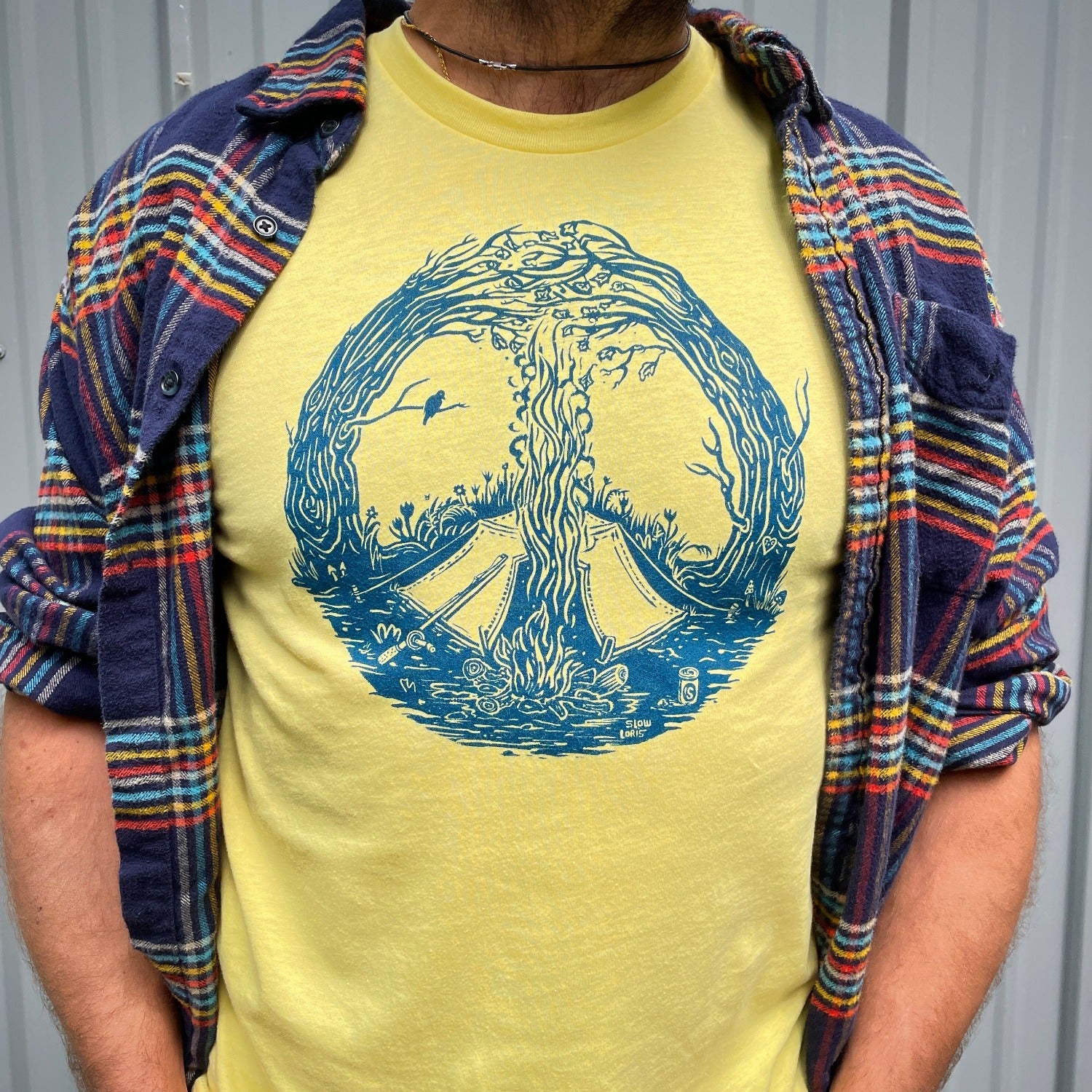  Close up of blue print on a yellow T. Print is of a campfire's smoke going up the center meeting with the tops of curved in trees creating a peace sign. Tent is by the fire and a bird is on a branch
