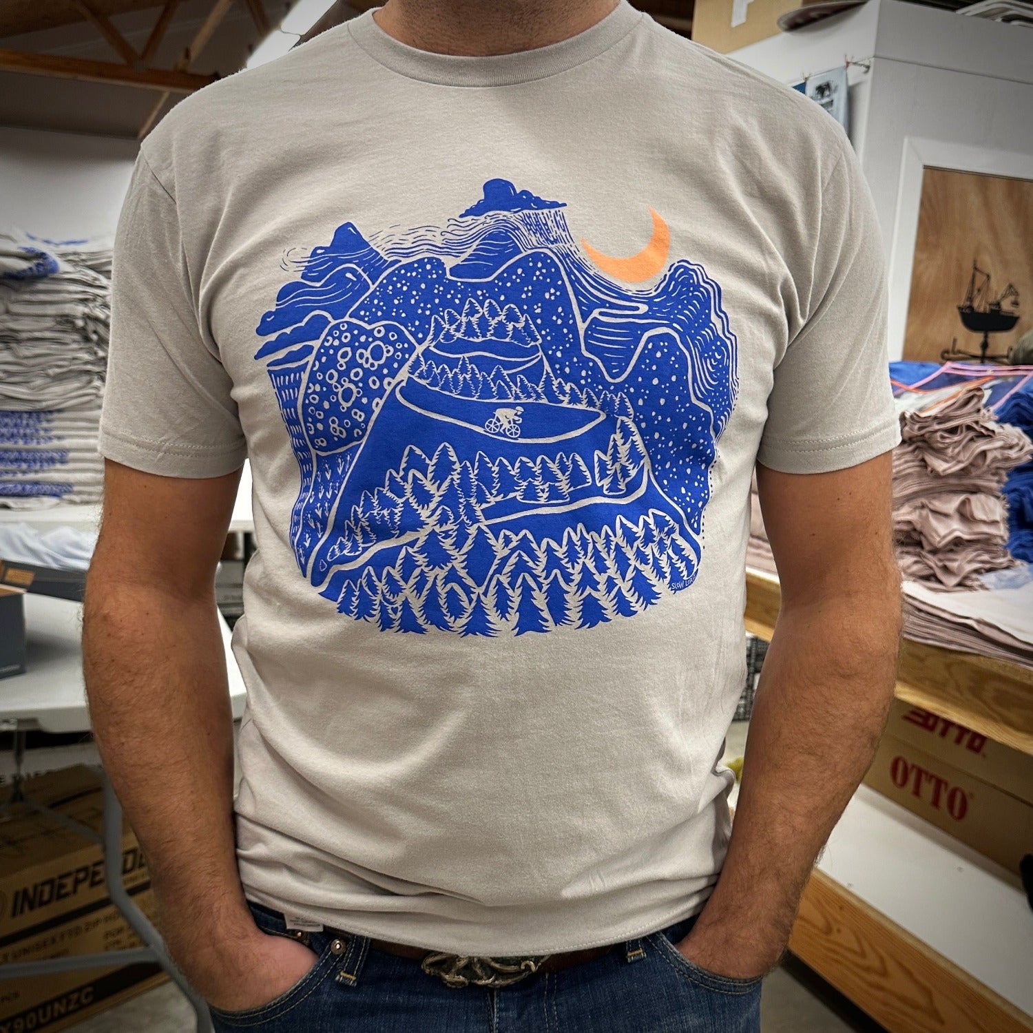 Night Ride print on tan shirt in dark blue ink. Mountain scene with raincloud, crescent moon, and bicyclist riding down the mountain