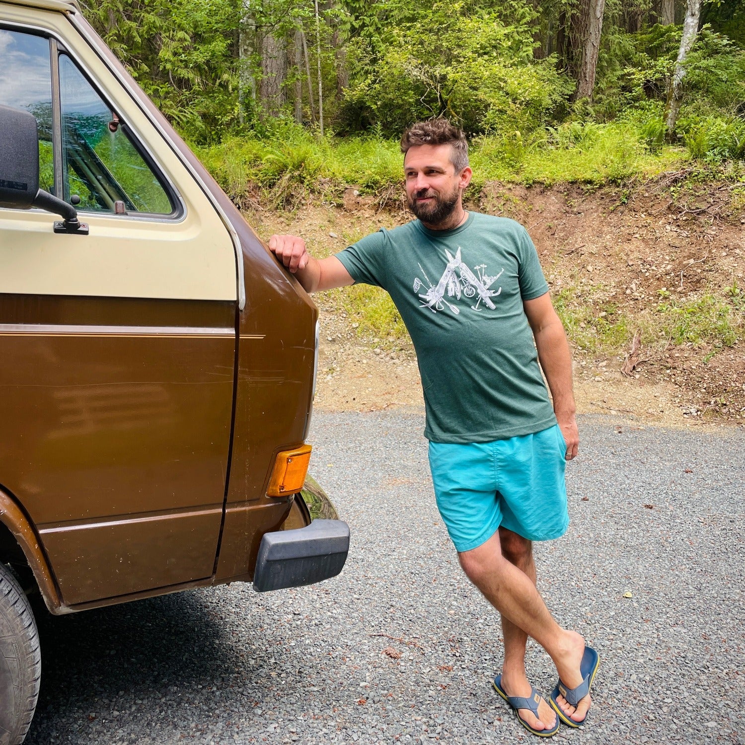 man wearing lighter green t-shirt with a white print of a multitool. The tools within the multitool are actually adventuring items- kayak, fishing pole, skis, ice axe, etc etc.