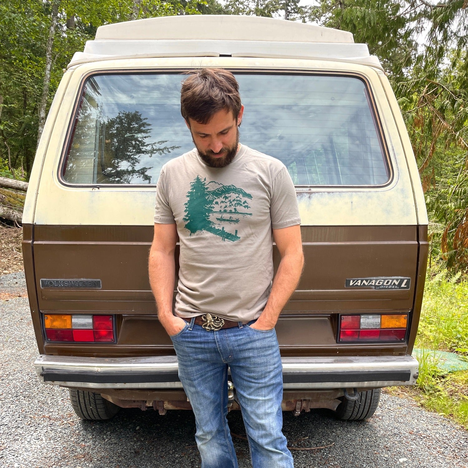 Man standing behind a VW can wearing a warm grey t-shirt with a green print of lakeside scene on it. 