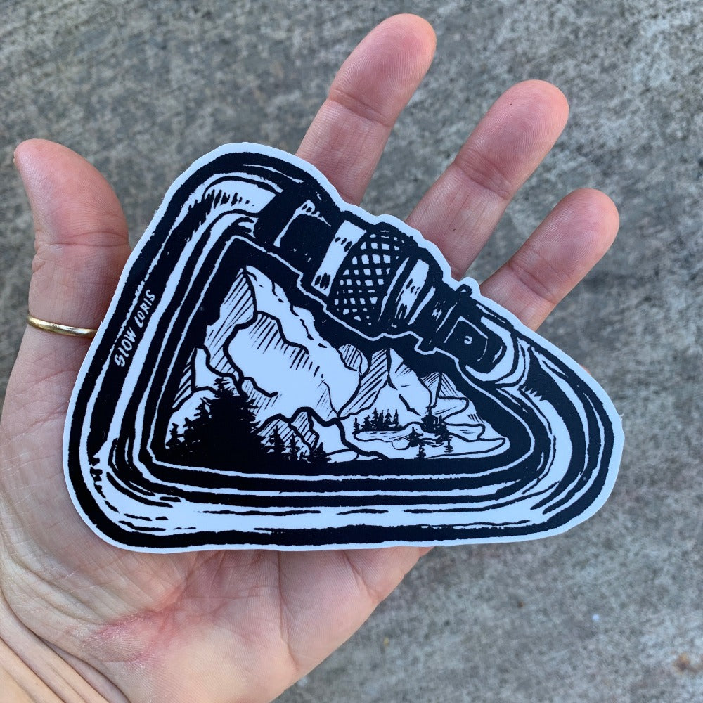 Black and white sticker of a mountain landscape framed by a carabiner. Sticker is held in the palm of a hand.