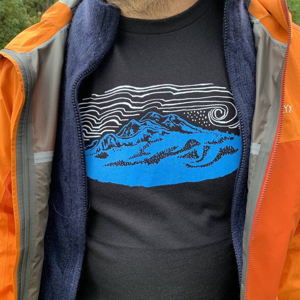 Black t shirt printed with a blue mountain and white skies, peeking out of an unzipped hoodie and jacket.
