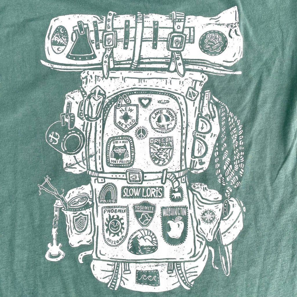 A screen printed design of an old-school backpack and bedroll covered in state and state park patches. The print is white on a green t shirt.