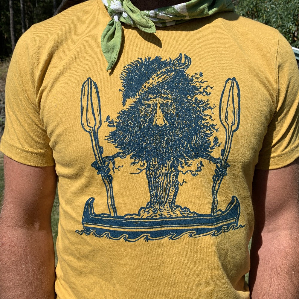T shirt printed with tree dude standing in a canoe, being worn by a man outside.