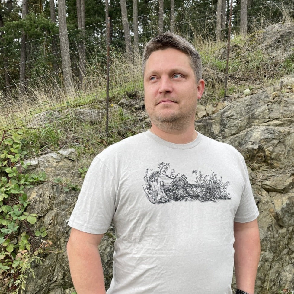 A man posing in a t shirt. The t shirt design is a guitar on its side, overgrown with nature, with bees around. Black print on a light gray tee. 