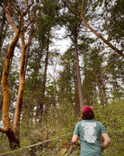 A man standing in the woods, facing away from the camera. He is wearing a t shirt screen printed with an old-school backpack and bedroll, covered in state and state park badges. The print is white on a green t.