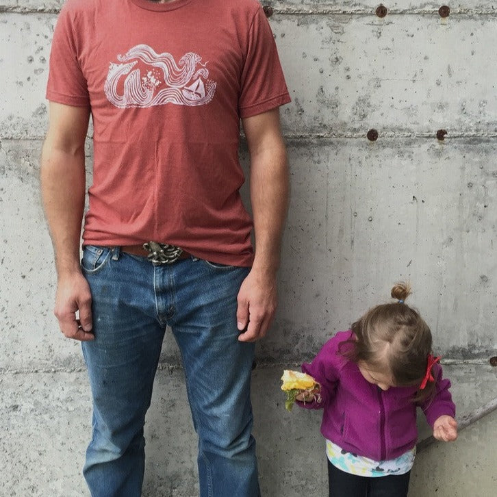 Man wearing clay t-shirt with white print of big winds and big seas tossing boats around. Next to model stands little toddler eating a huge sandwich